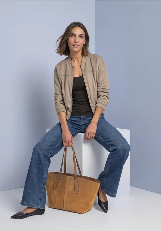 Frau sitzt auf weißem Würfel, trägt beige Jacke, schwarzen Top, weite Jeans und hält braune Tasche.
