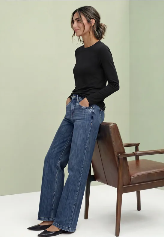 Junge Frau steht in weitem Jeans und schwarzem Langarmshirt neben einem braunen Stuhl.