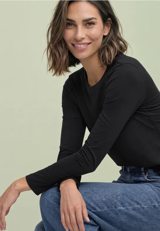 Eine lächelnde Frau in schwarzem Langarmshirt und Jeans sitzt lässig und lehnt sich vor.