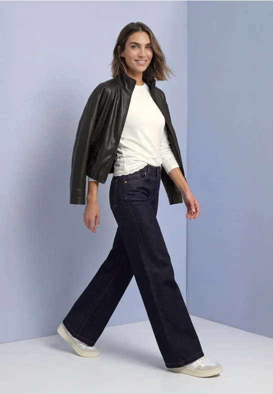 Frau mit weitem Jeans, weißem Shirt und Lederjacke über der Schulter, die selbstbewusst lächelnd geht.