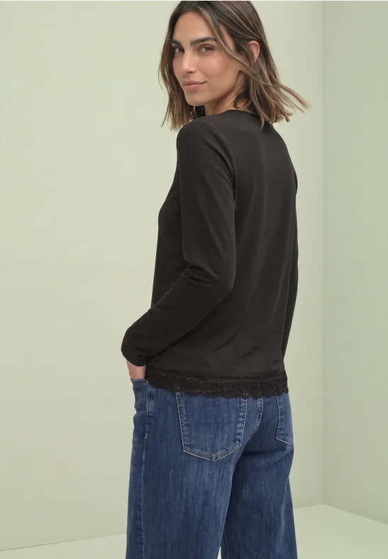 Frau mit schwarzen Langarmshirt und weiten Jeans, halb im Profil, vor hellem Hintergrund.