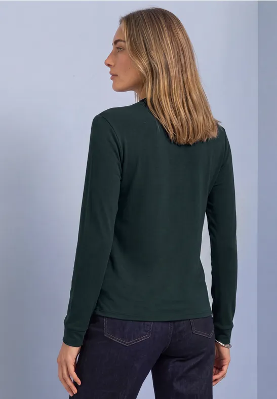 Vrouw van achteren in een donkergroen shirt met lange mouwen en een spijkerbroek voor een neutrale achtergrond.