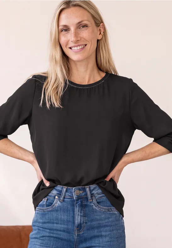 Lächelnde Frau mit schwarzem T-Shirt und Jeans, steht mit Händen in der Hüfte vor neutralem Hintergrund.