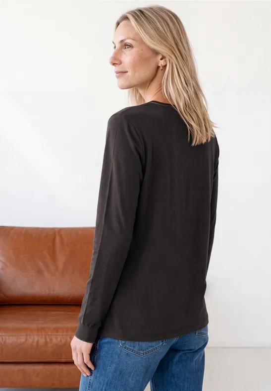 Blonde Frau trägt schwarzes Langarmshirt und blaue Jeans, steht seitlich vor einem braunen Sofa, Blick nach links.