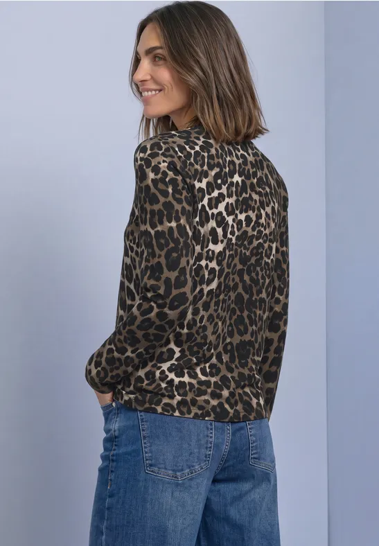 Lächelnde Frau mit schulterlangem Haar trägt Leoprintbluse mit langen Ärmeln und Jeans vor grauem Hintergrund.