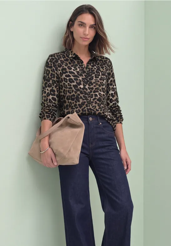 Eine Frau vor grünem Hintergrund trägt ein Leopardenmuster-Hemd, weite Jeans und hält eine beige Tasche.
