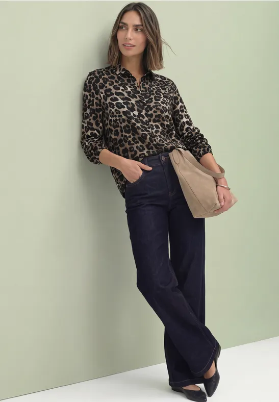 Frau lehnt an grüner Wand, trägt Leopardenbluse, weite Jeans, schwarze Schuhe und eine Beuteltasche.