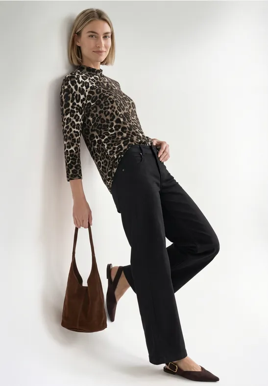Frau lehnt an weißer Wand, trägt Leopardenprint-Top, schwarze Hose, braune Tasche und Loafers.