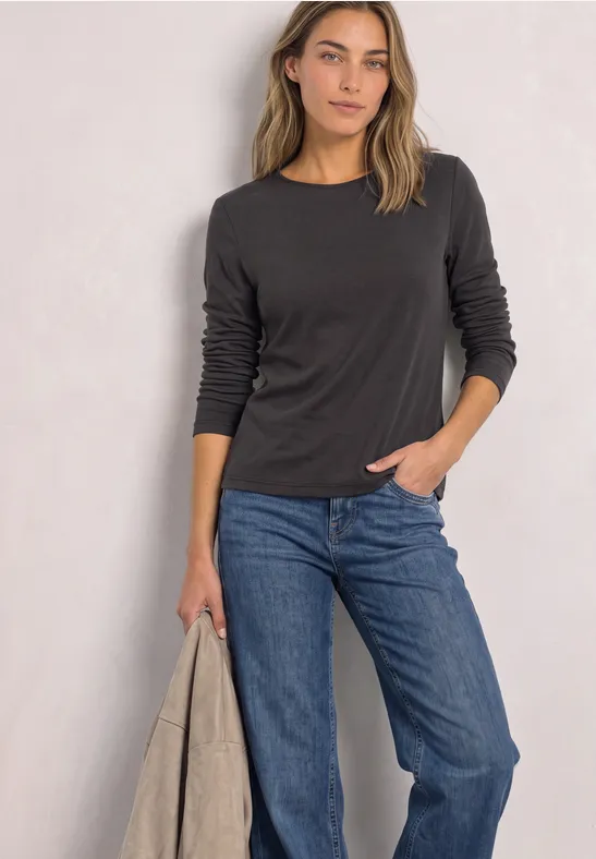Frau mit schwarzem Langarmshirt, blauen Jeans und heller Jacke in der Hand vor hellem Hintergrund.