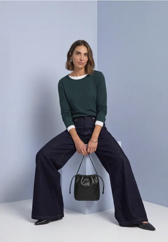 Vrouw in donkergroene trui, wijde broek en zwarte schoenen zit, houdt een zwarte handtas vast.