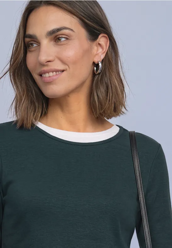 Een glimlachende vrouw met halflang bruin haar, groen shirt met witte kraag en een tas over de schouder.