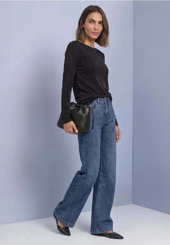 Frau in schwarzem Oberteil, weitem Jeans, spitzen Schuhen, hält eine schwarze Clutch vor hellgrauer Wand.
