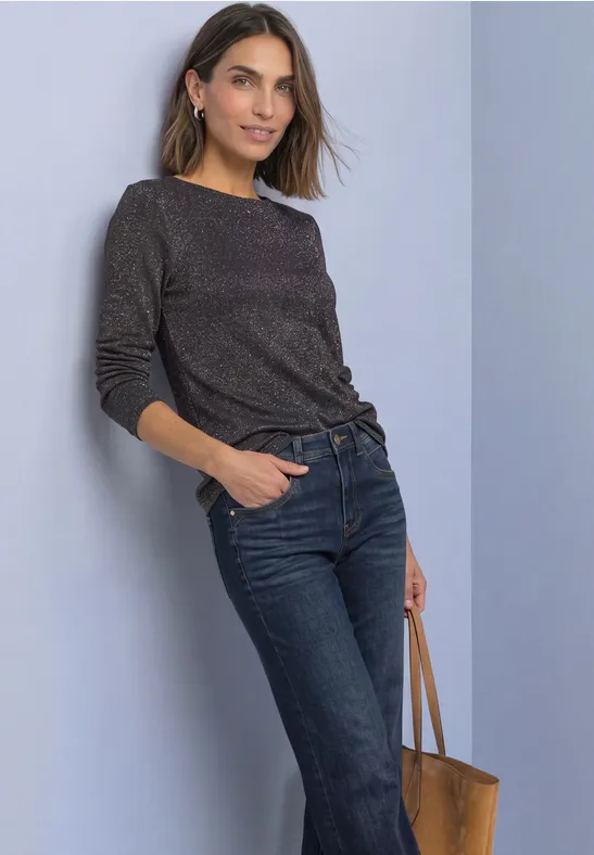 Vrouw leunt tegen muur, zwarte trui, blauwe jeans en bruine leren tas.