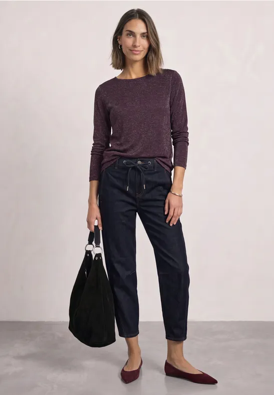 Frau trägt lila Pullover, hohe Jeans und burgunderfarbene Ballerinas, hält schwarze Tasche.