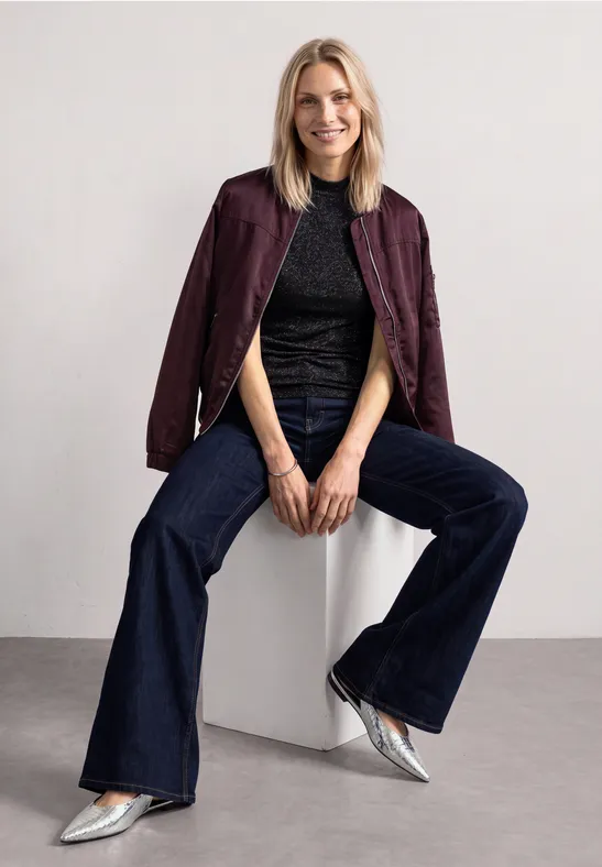Eine Frau sitzt auf einem weißen Block, trägt burgunderfarbene Jacke, schwarzes Top, weite Jeans und silberne Schuhe.