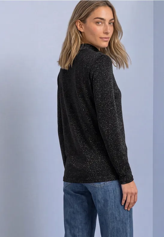 Frau in schwarzem Glitzerpullover und blauer Jeans blickt über die Schulter.