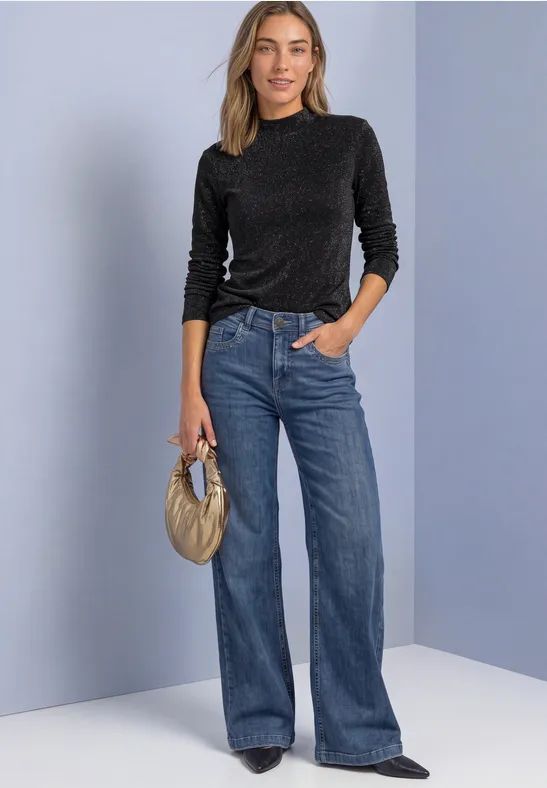 Frau in schwarzem Pullover und blauen Jeans hält goldene Handtasche vor neutralem Hintergrund.