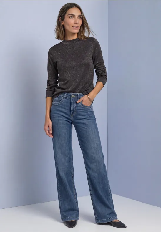 Frau in schwarzem Langarmshirt, weiten blauen Jeans und schwarzen Schuhen vor lila Hintergrund.