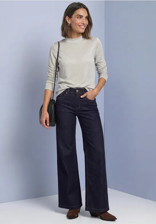 Frau trägt hellgrauen Pullover, hoch taillierte, weite Jeans, schwarze Umhängetasche und braune Schuhe.