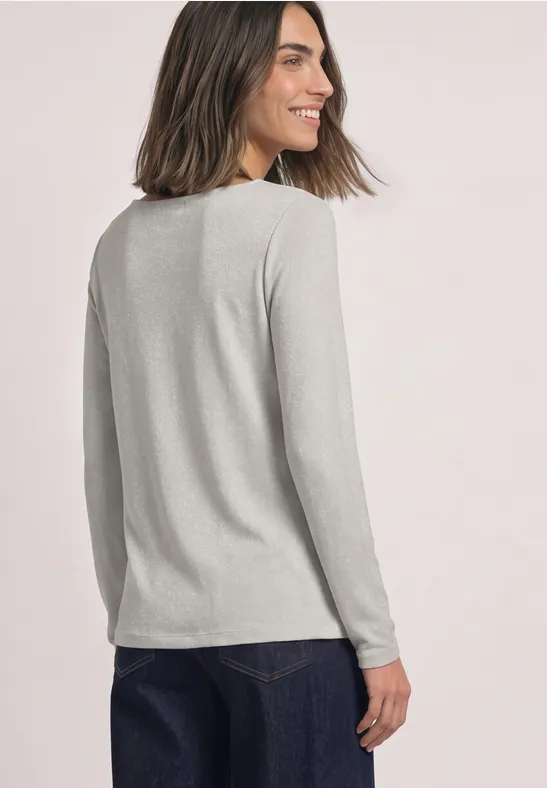 Frau leicht seitlich von hinten, lächelt, trägt graues Langarmshirt und Jeans