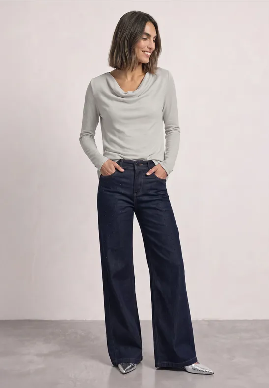 Frau in weißem Wasserfallkragenoberteil und weiten dunkelblauen Jeans, Hände in den Taschen, silberne Schuhe.