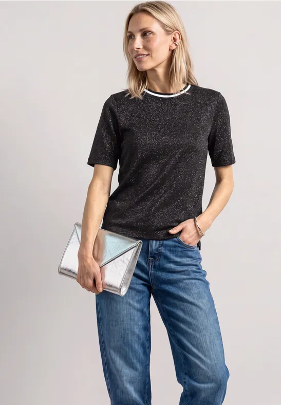 Frau in schwarzem Glitzer-Shirt, blauer Jeanshose und silberner Clutch vor neutralem Hintergrund.