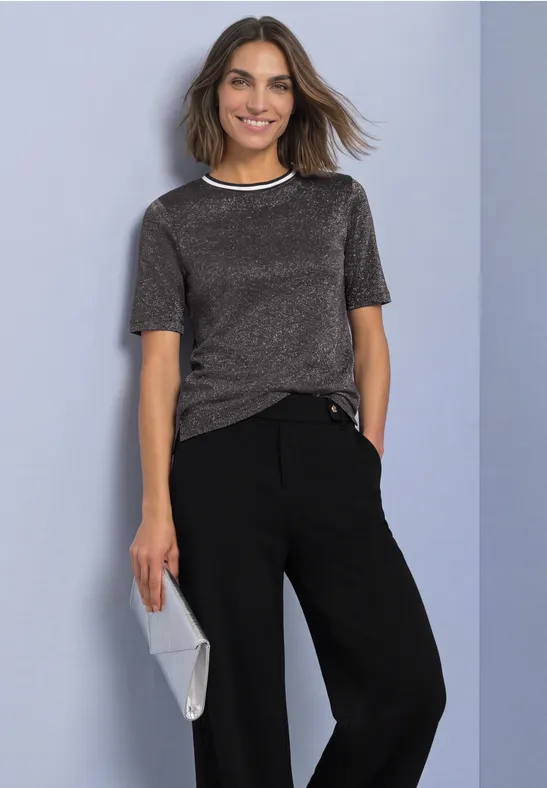 Een glimlachende vrouw voor een grijsblauwe achtergrond, in een zilverkleurig T-shirt, zwarte broek en grijze clutch.