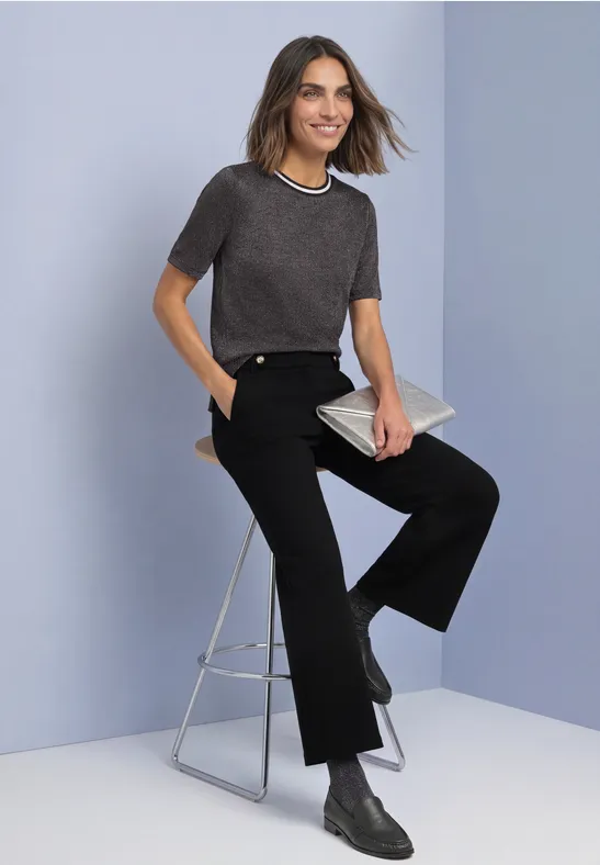 Vrouw in grijs shirt en zwarte broek zit op een kruk, houdt een clutch vast en draagt enkellaarzen.