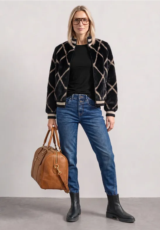 Femme en bomber à motif, pull noir, jean bleu et bottines noires, tenant un sac en cuir marron.