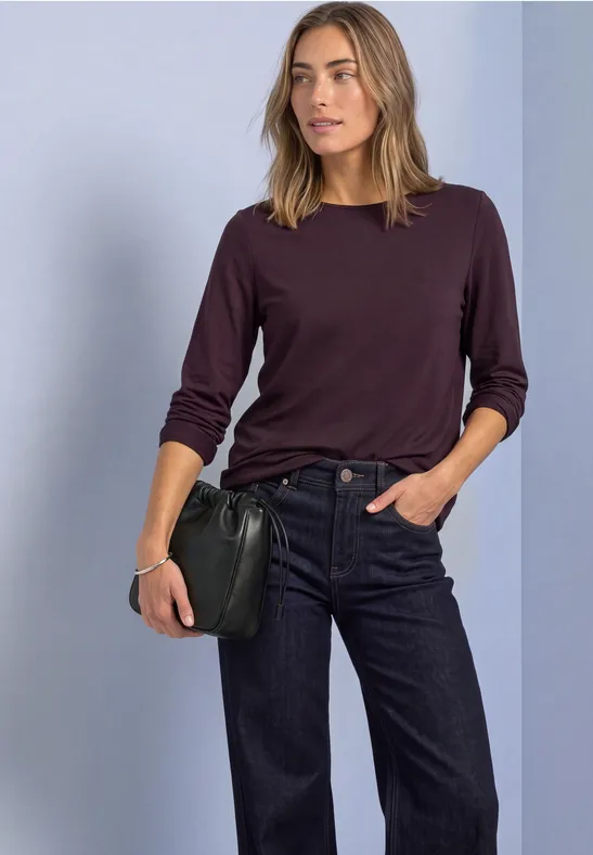 Vrouw in donkerblauwe jeans en bordeaux top met lange mouwen, houdt zwarte clutch vast voor lichte achtergrond.