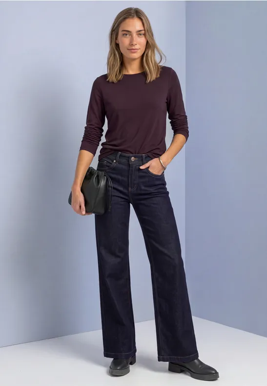 Vrouw in donkerrood trui, zwarte wijde jeans en schoenen, met zwarte schoudertas, voor grijs-paarse achtergrond.