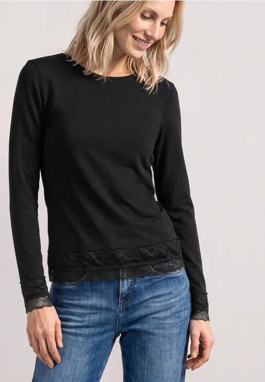 Frau in schwarzem Langarmshirt mit Spitzenbesatz an Bündchen und Saum, dazu Blue Jeans, lächelnd