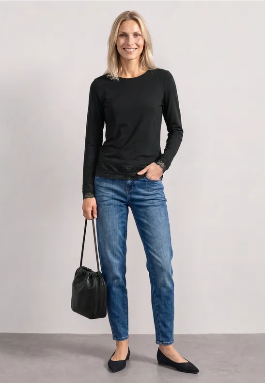 Frau steht vor hellgrauem Hintergrund, trägt schwarzen Pullover, Jeans, flache Schuhe und hält eine schwarze Tasche.