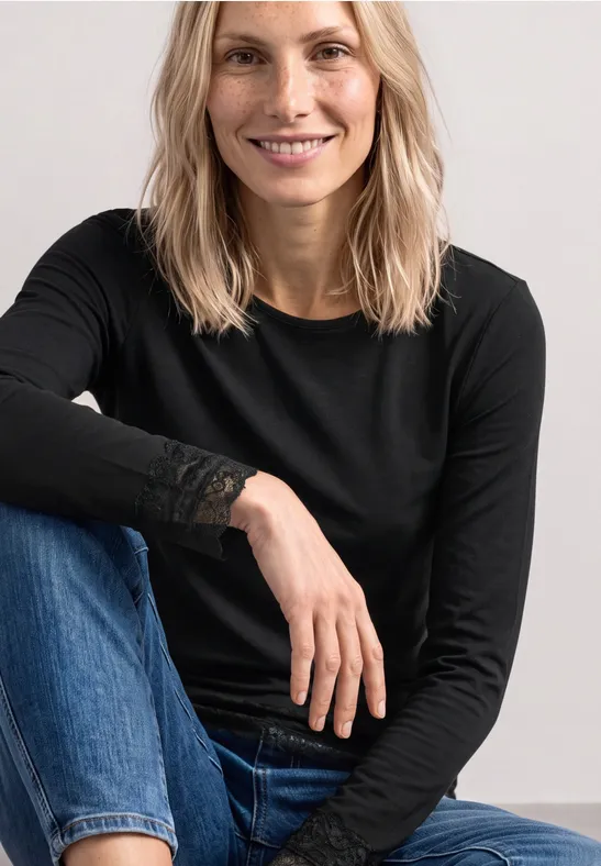Eine blonde Frau sitzt lächelnd trägt schwarzen Langarmshirt mit Spitzendetails und blaue Jeans vor schlichten Hintergrund.