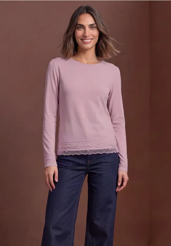 Vrouw lacht in een roze longsleeve met kanten rand en blauwe jeans voor een bruine achtergrond.