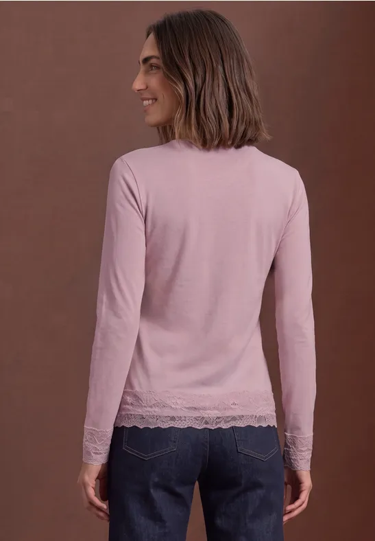 Een vrouw in een lichtroze top met kant en een spijkerbroek staat opzij voor een bruine achtergrond.