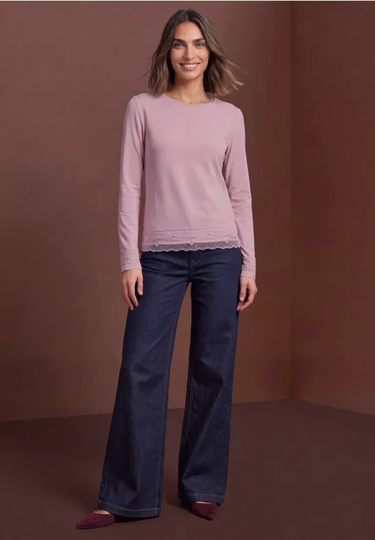 Vrouw in roze longsleeve met kant, wijde spijkerbroek en rode schoenen voor een bruine achtergrond.