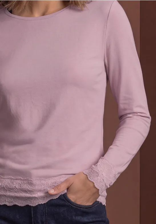 Vrouw draagt lichtpaars shirt met lange mouwen en kantafwerking, hand in de zak.