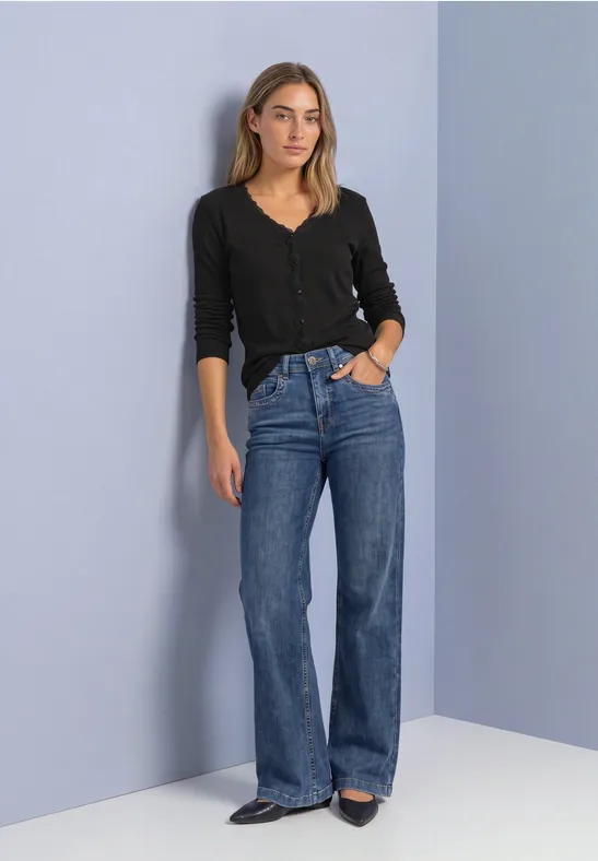 Eine Frau steht lässig vor einer hellen Wand, trägt schwarzen Pullover und gerade geschnittene blaue Jeans.
