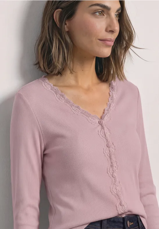 Frau mit schulterlangem braunem Haar, die ein hellrosa Spitzen-Cardigan mit V-Ausschnitt trägt und seitlich blickt.