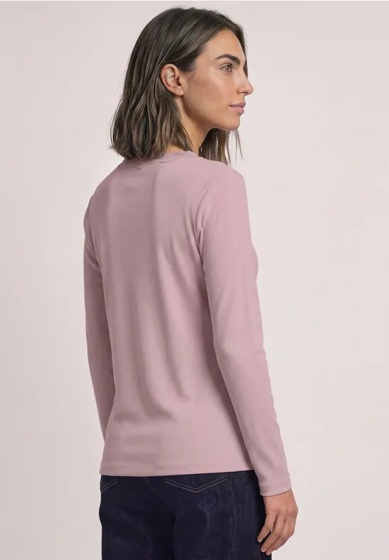 Eine Frau von hinten in seitlicher Profilansicht trägt ein rosa Langarmshirt und dunkle Jeans vor hellem Hintergrund.