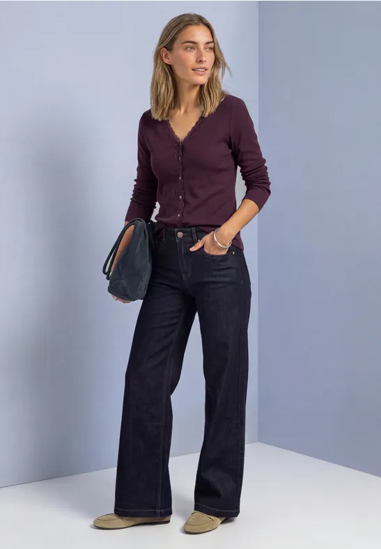 Eine Frau steht vor hellvioletter Wand, trägt eine bordeauxfarbene Strickjacke, weite Jeans und hält eine Clutch.
