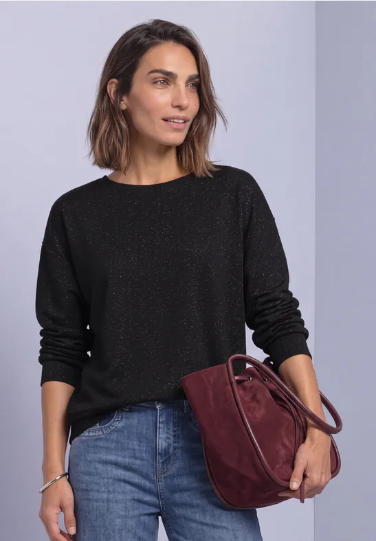 Eine Frau trägt einen schwarzen Pullover, Jeans und eine dunkelrote Tasche vor hellem Hintergrund.