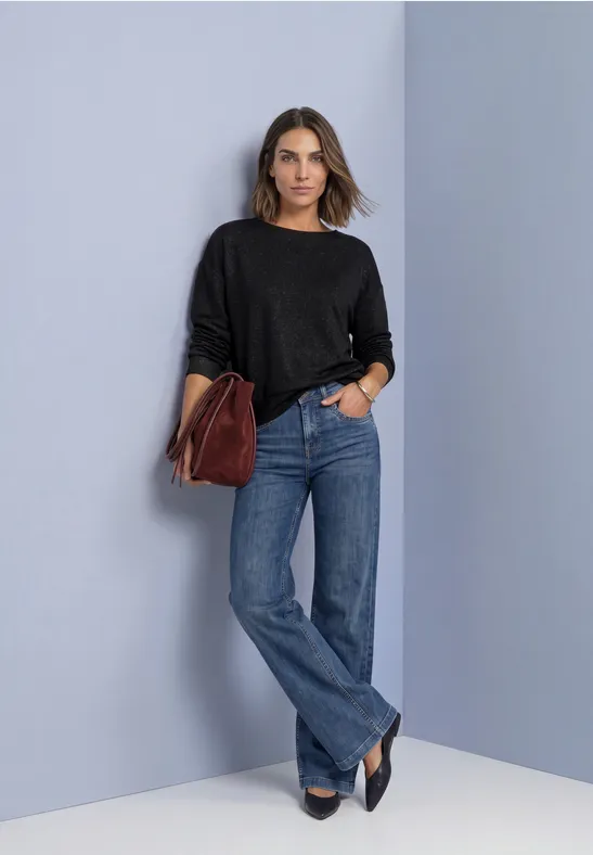 Frau steht vor hellgrauer Wand, trägt schwarzen Pullover, gerade geschnittene Jeans, schwarze Schuhe und rote Tasche.