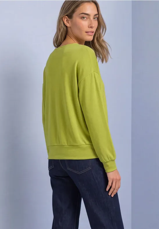 Profilaufnahme einer Frau in grünem Pullover und blauer Jeans vor hellem Hintergrund.