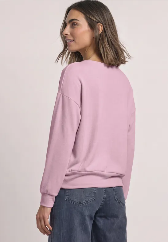Frau steht seitlich, trägt rosafarbenen Pullover und blaue Jeans, kurz geschnittenes Haar, weißer Hintergrund.