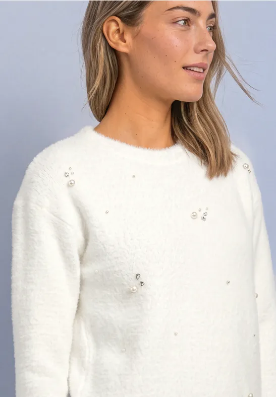 Junge Frau in weißem Strickpullover mit dezenten Applikationen vor grauem Hintergrund.