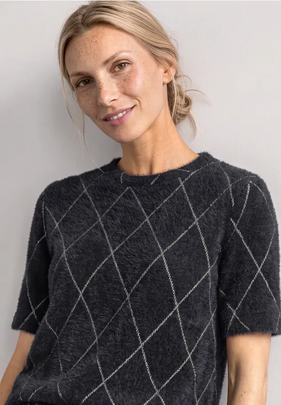 Frau im schwarzen Strickshirt mit Rautenmuster, lächelnd vor hellem Hintergrund.