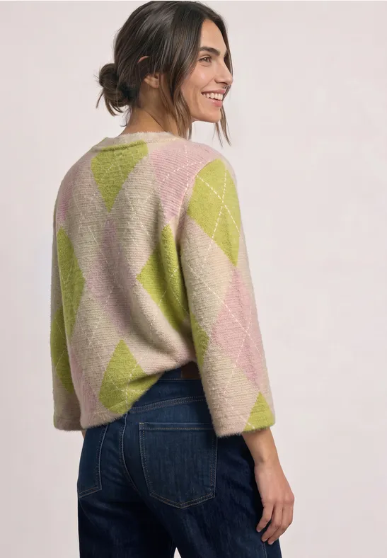 Frau im Profil trägt bunte Argyle-Strickjacke in Pastelltönen und Jeans, lächelnd vor hellem Hintergrund.