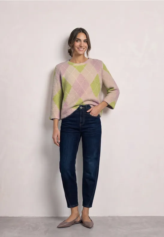 Frau in pastellgemustertem Pullover und dunklen Jeans, lächelnd und entspannt vor heller Wand stehend.